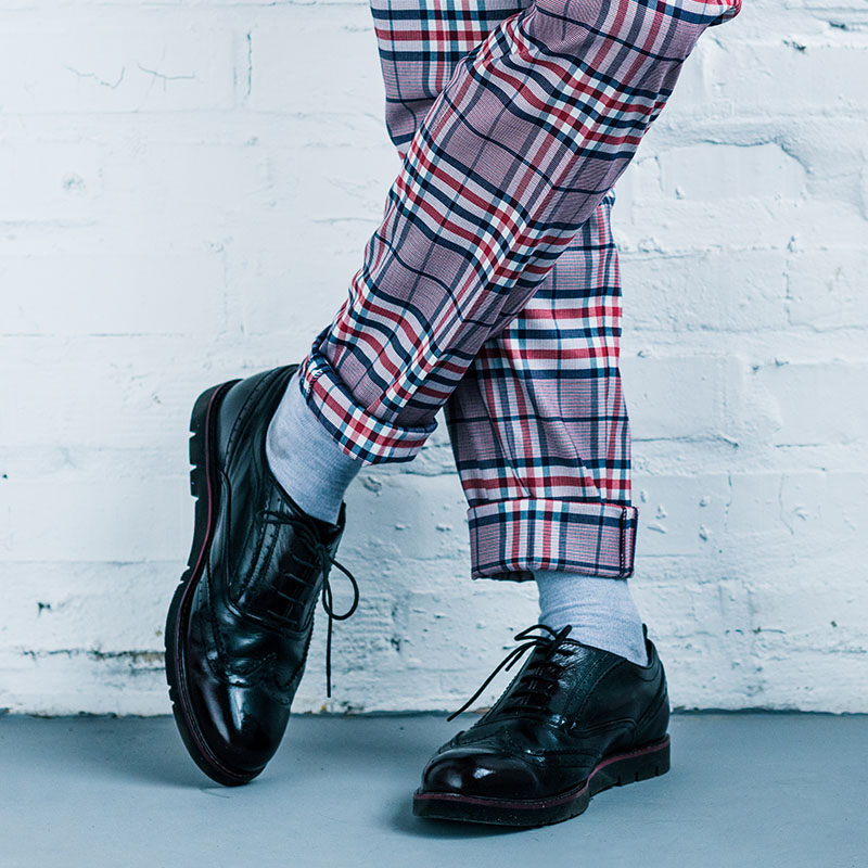 Gambe di uomo che indossano pantalone in tartan, calzini grigi e scarpe eleganti nere. Sullo sfondo un muro bianco e un pavimento grigio